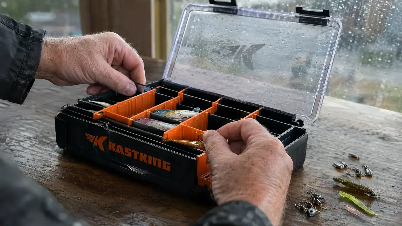 Hands sorting hooks, swivels, and lures into the labeled compartments of an open KastKing tackle tray at a kitchen table
